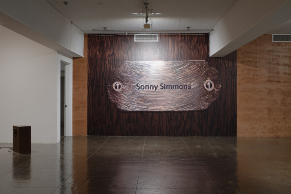 “DON'T LET 'EM PICK GUITARS AND DRIVE THEM OLD TRUCKS, MAKE 'EM BE DOCTORS AND LAWYERS AND SUCH”<br/>
2011 ongoing <br/>
Wall: C-Print Wall Mural of an Imaginary Name Plate Reserving a Hotel Room in Perpetuity for the Jazz Legend, Sonny Simmons, Mounted between Wood Paneling<br/>
Variable Dimensions<br/>
Floor: 1 Kenwood KL777 Speaker Radiating the Spoken Word Piece, “Sonny Simmons Comments on 56 Jazz Legends” in a Continuous Loop<br/>
Shipping Box: Variable Dimensions<br/>
Speakers: 16” x 25” x 11 1/2”<br/>